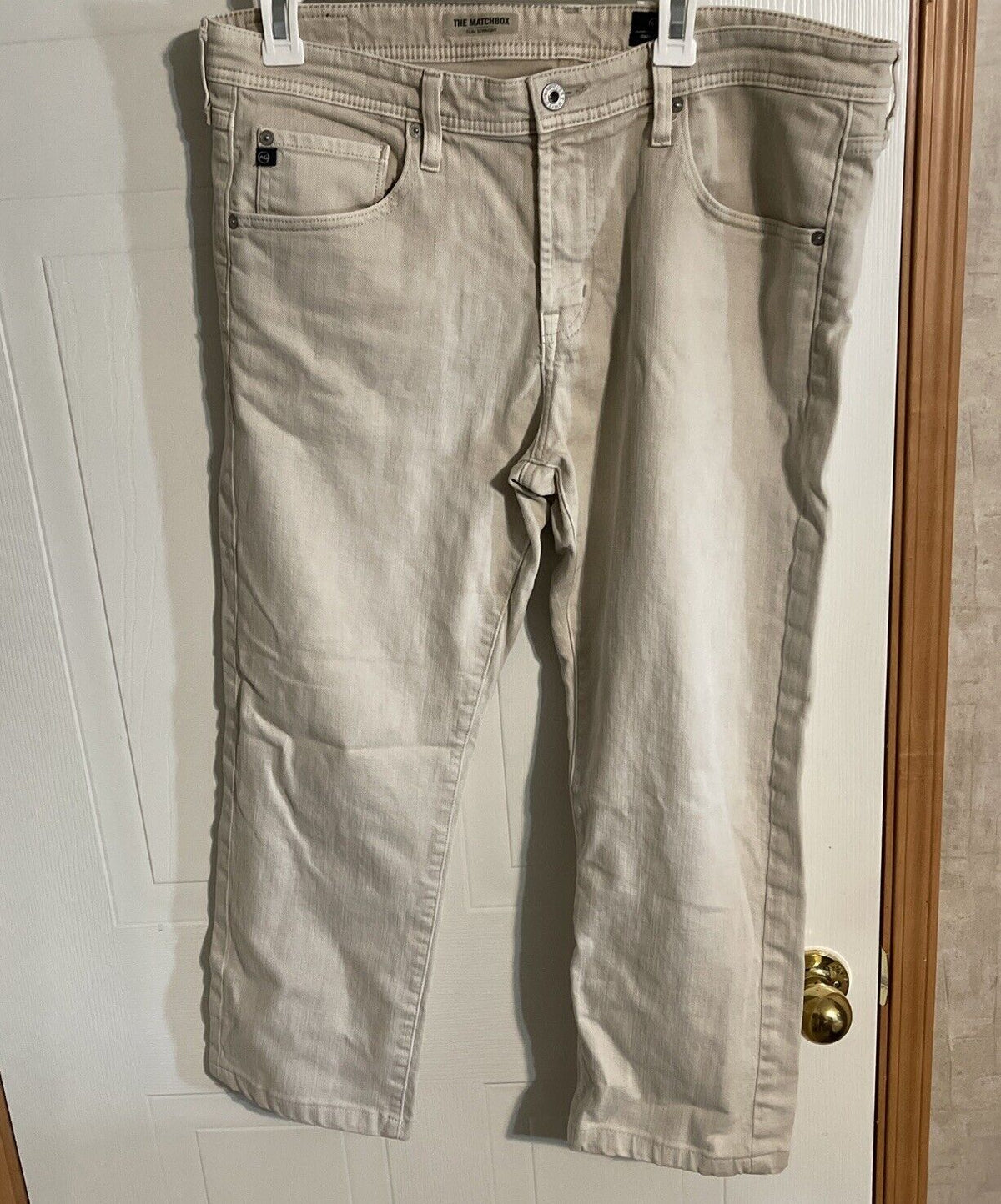 Beige Adriano Goldschmied Matchbox jeans hanging on white door with front pockets and button closure