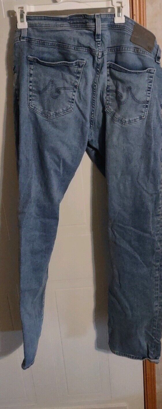 AG Adriano Goldschmied jeans men in light blue denim with back pockets and belt loops hanging on a hanger