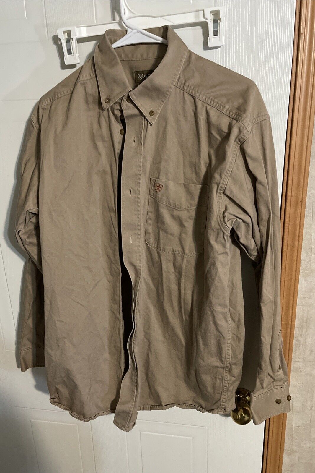 Ariat men’s button down shirt in tan hanging on white plastic hanger with chest pocket and long sleeves