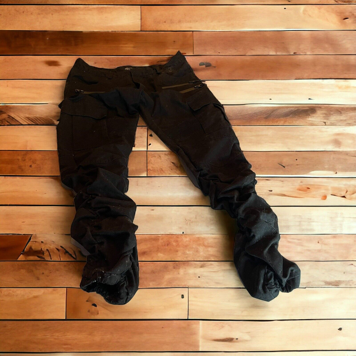 Black cargo pants with zipper pockets laid flat on a wooden floor showing front and side details